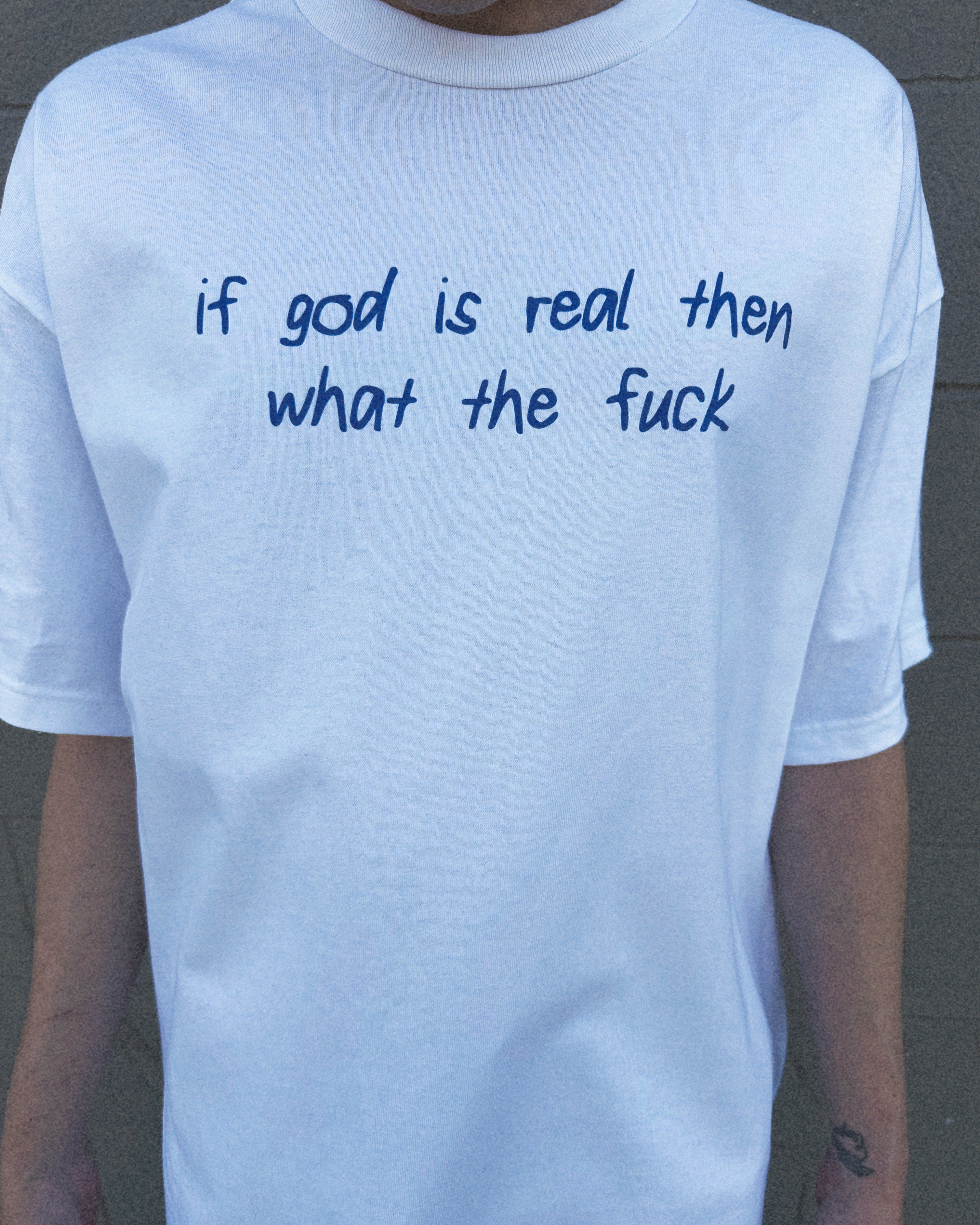 White t-shirt with blue text 'if god is real then what the fuck' worn by a person.