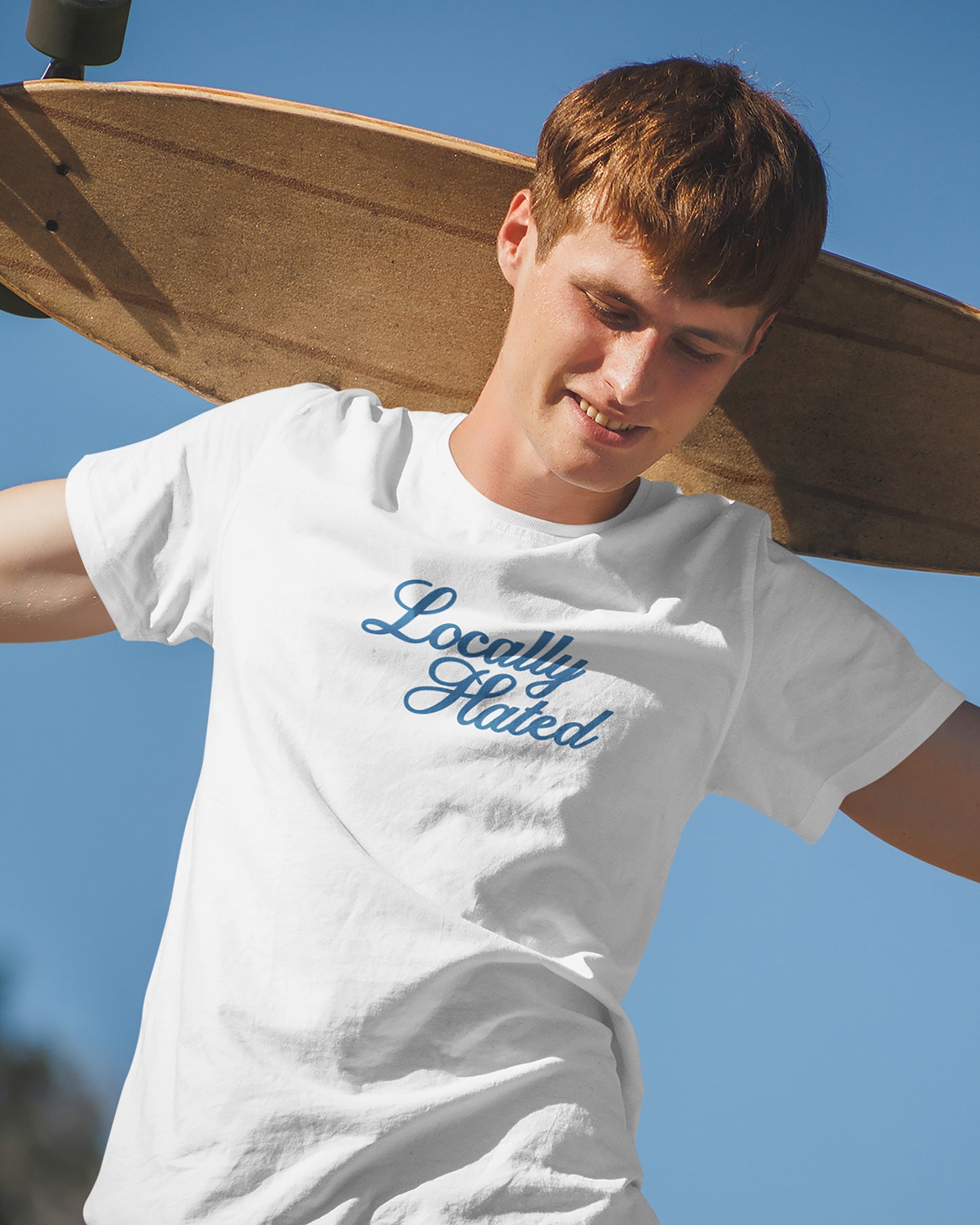 Person wearing a white t-shirt with 'Locally Hated' text, holding a skateboard against a clear blue sky.