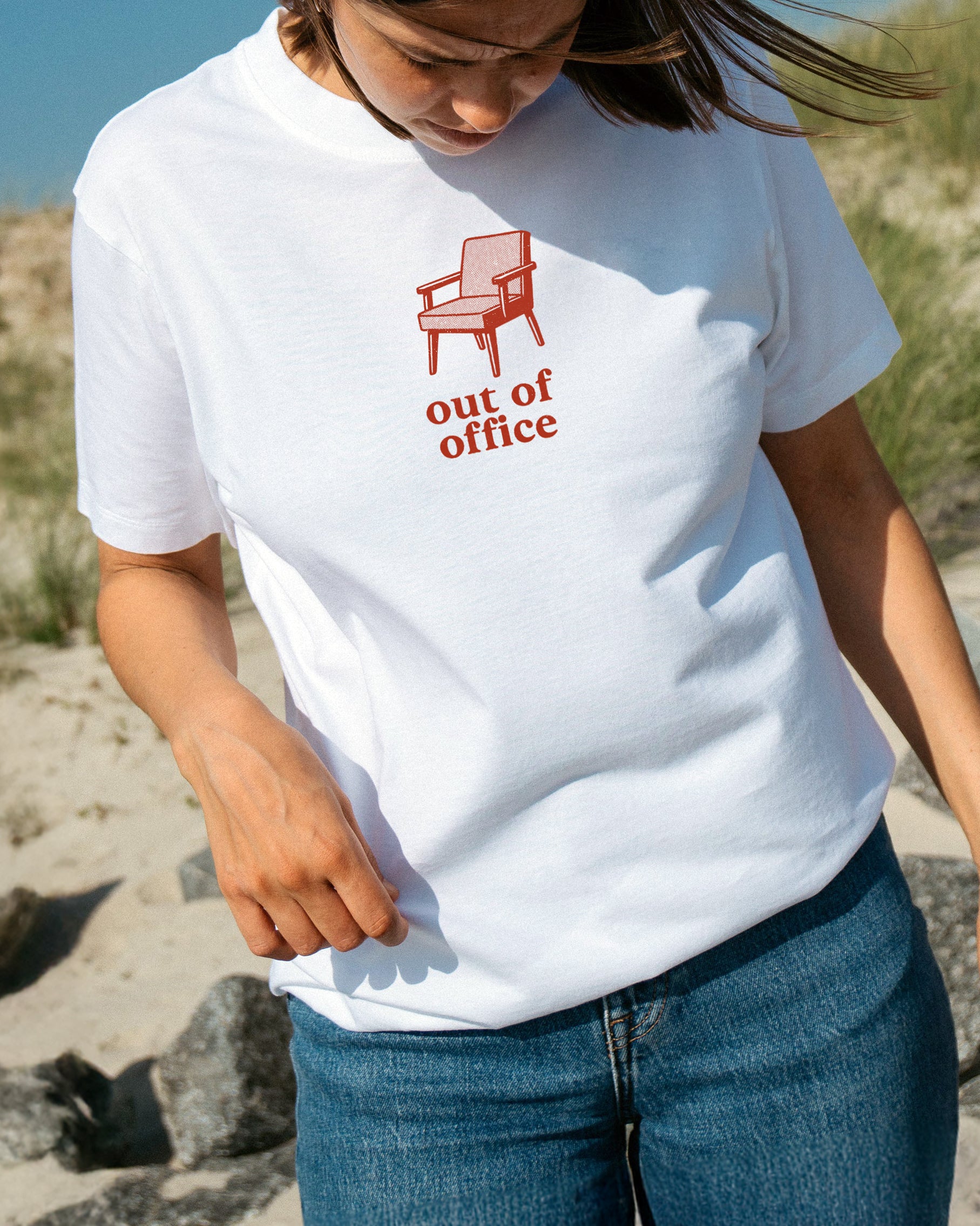Person wearing a white t-shirt with a red chair graphic and 'out of office' text on a beach.