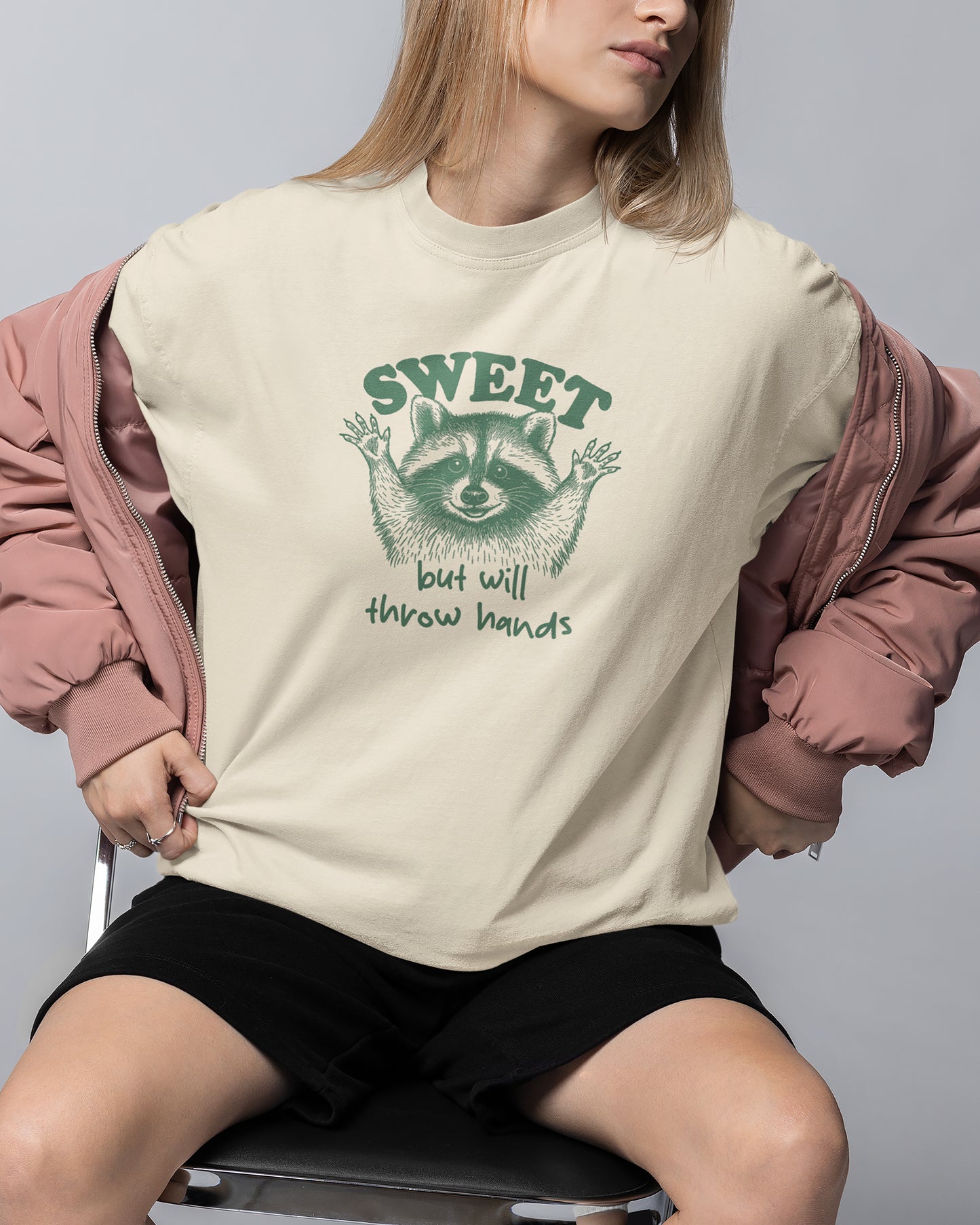 Person wearing a cream t-shirt with a raccoon graphic and text, sitting on a chair against a gray background.