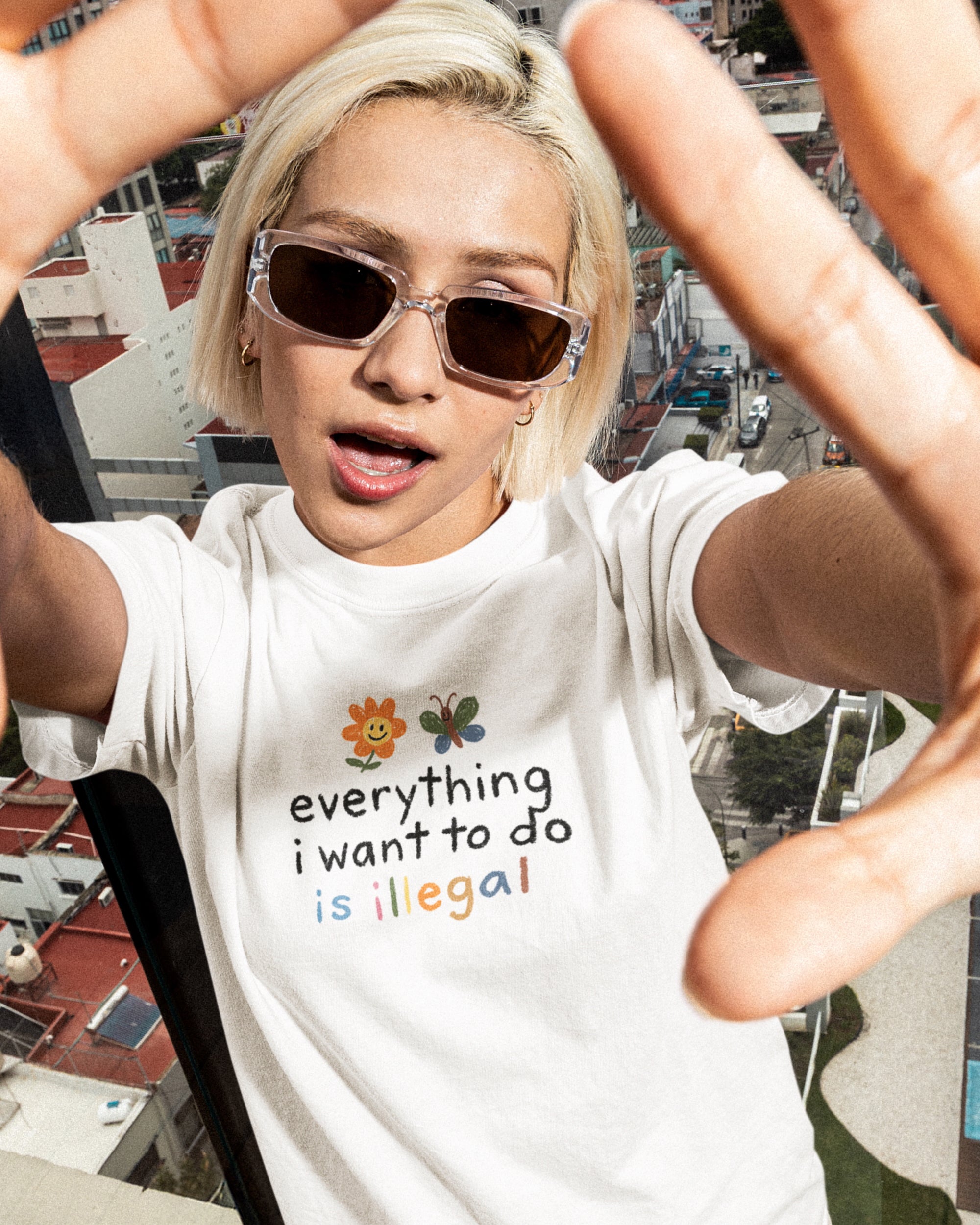 Person wearing a white t-shirt with a "everything i want to do is illegal" design, taking a selfie with a cityscape background.