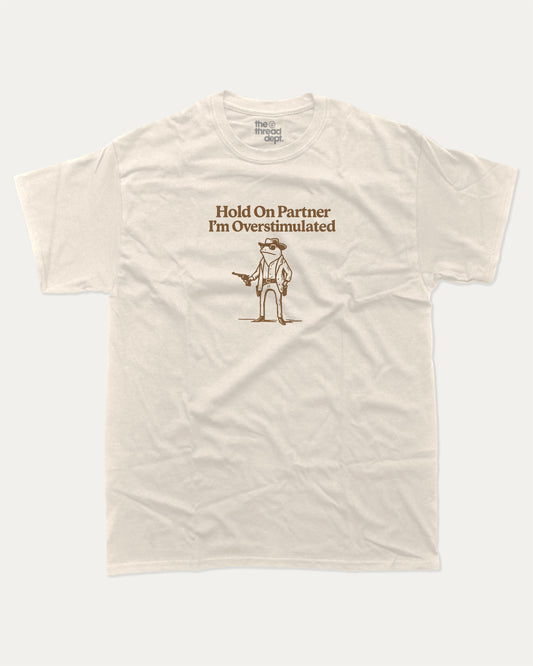 Hold On Partner I'm Overstimulated graphic t-shirt, perfect for adults. Funny Frogs tee, trending meme shirt, unique gift idea. Cream.