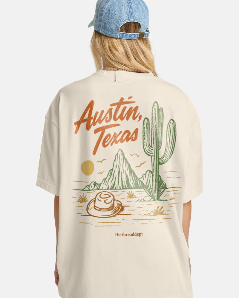 Person wearing a beige t-shirt with 'Austin, Texas' and desert graphics, standing against a white background.