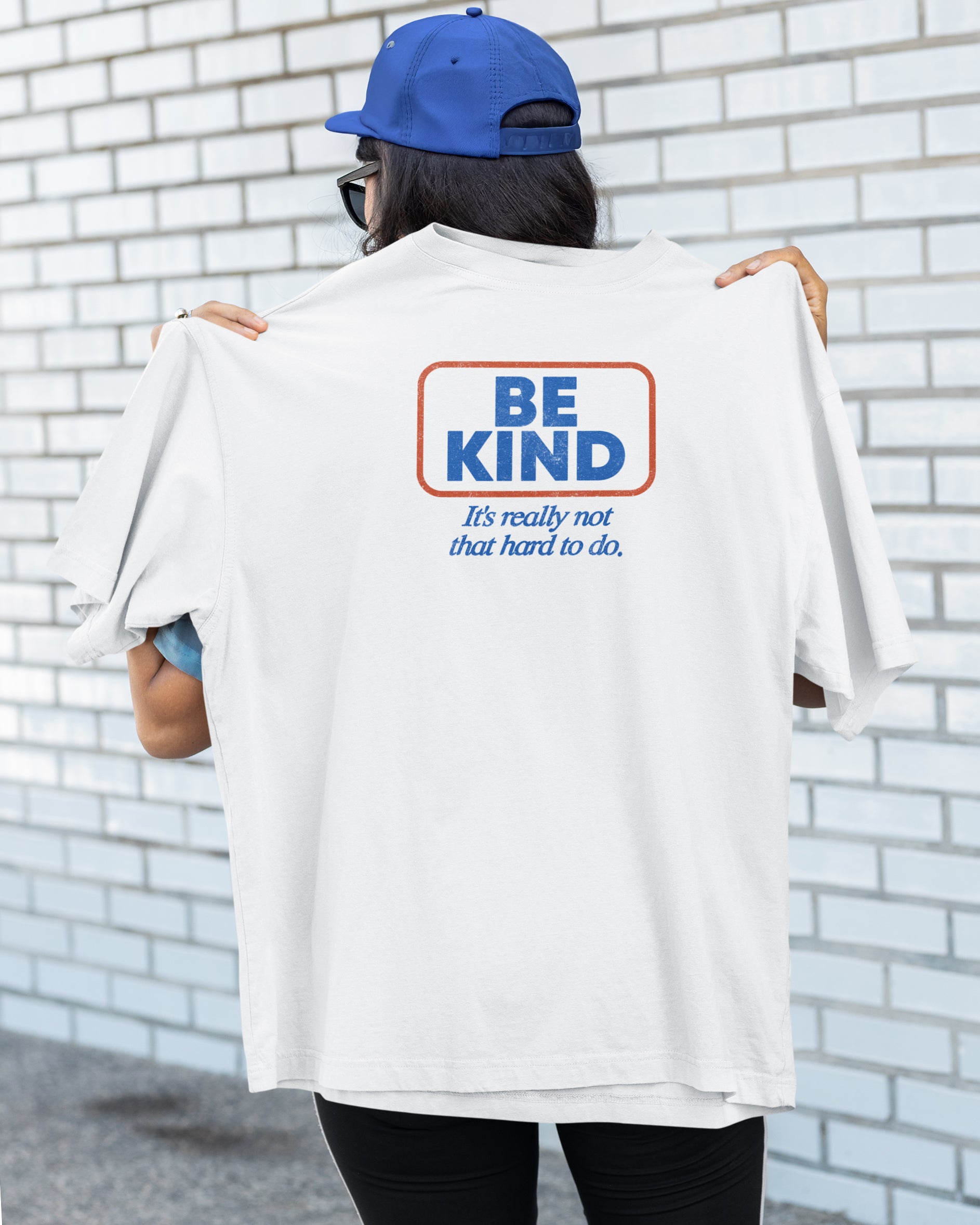 Person wearing a white t-shirt with 'Be Kind' message against a brick wall.