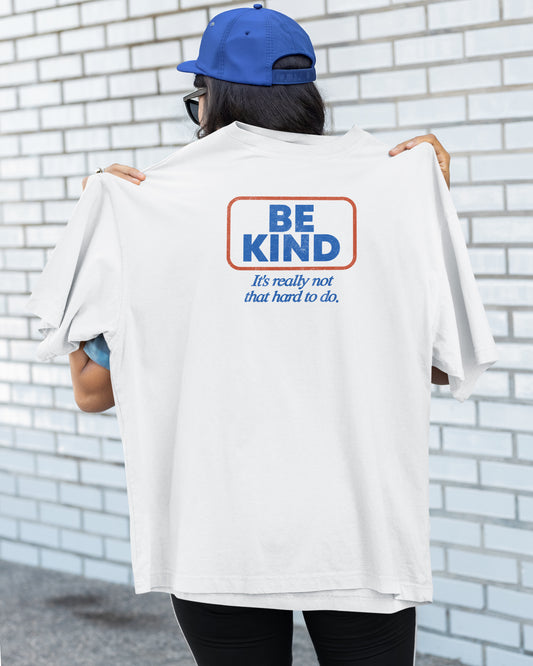 Person wearing a white t-shirt with 'Be Kind' message against a brick wall.