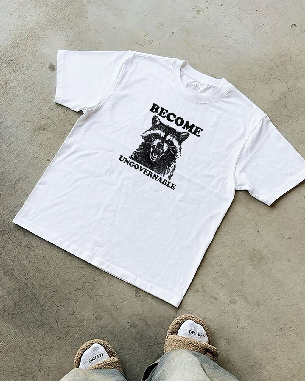 White t-shirt with a graphic of a raccoon and "become ungovernable" text on a concrete floor.