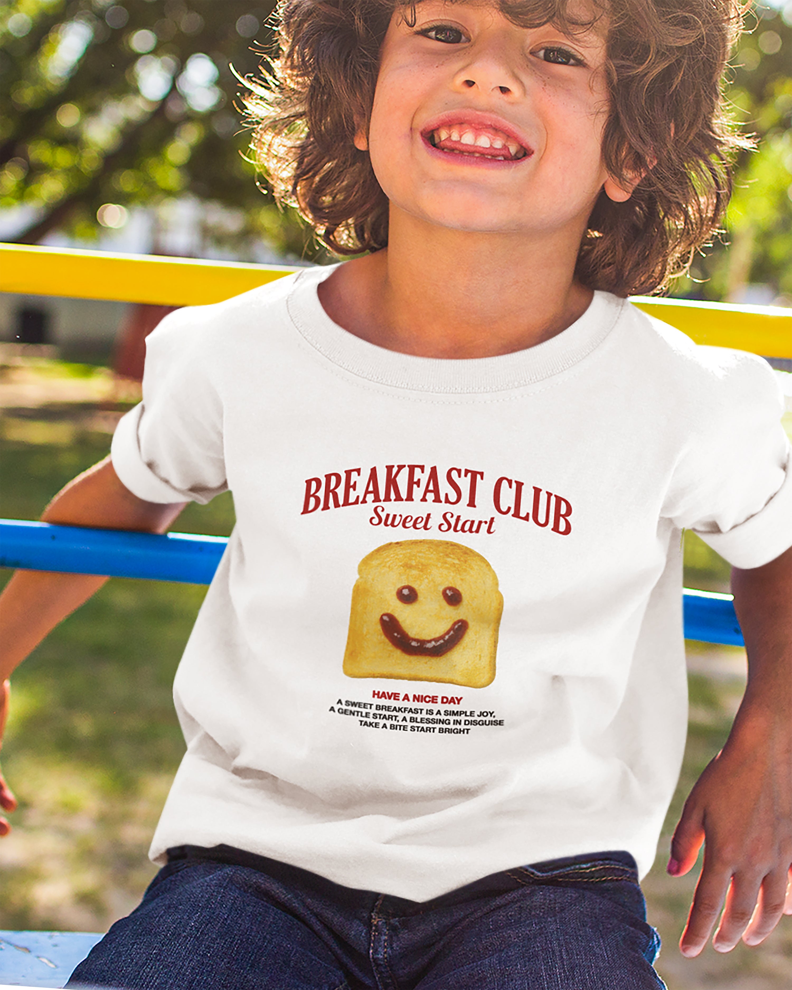 Child wearing a 'Breakfast Club Sweet Start' t-shirt with a smiley face toast graphic outdoors.