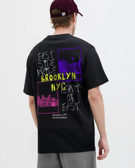 Person wearing a black t-shirt with colorful text and brooklyn nyc graphics on the back, standing against a white background.