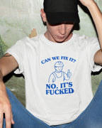 Person wearing a white t-shirt with "can we fix it no it's fucked" text and graphic, sitting against a textured wall.