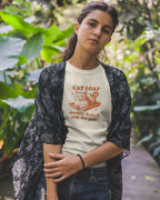 Woman wearing a t-shirt with a cat graphic and text, standing outdoors among greenery.