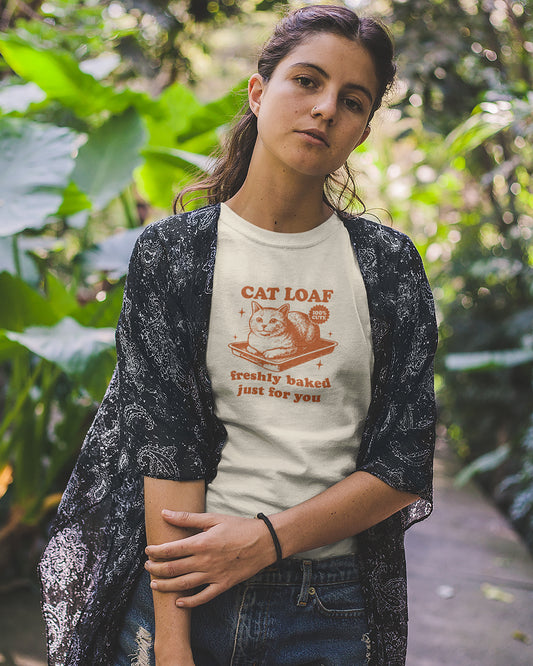 Woman wearing a t-shirt with a cat graphic and text, standing outdoors among greenery.