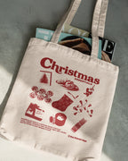 Beige tote bag with red Christmas illustrations and text on a gray surface