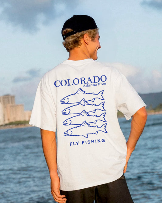 Person wearing a white t-shirt with 'colorado fly fishing' graphics and text by a body of water.