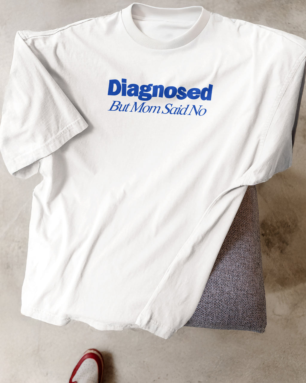 White t-shirt with blue text 'Diagnosed But Mom Said No' on a concrete floor.