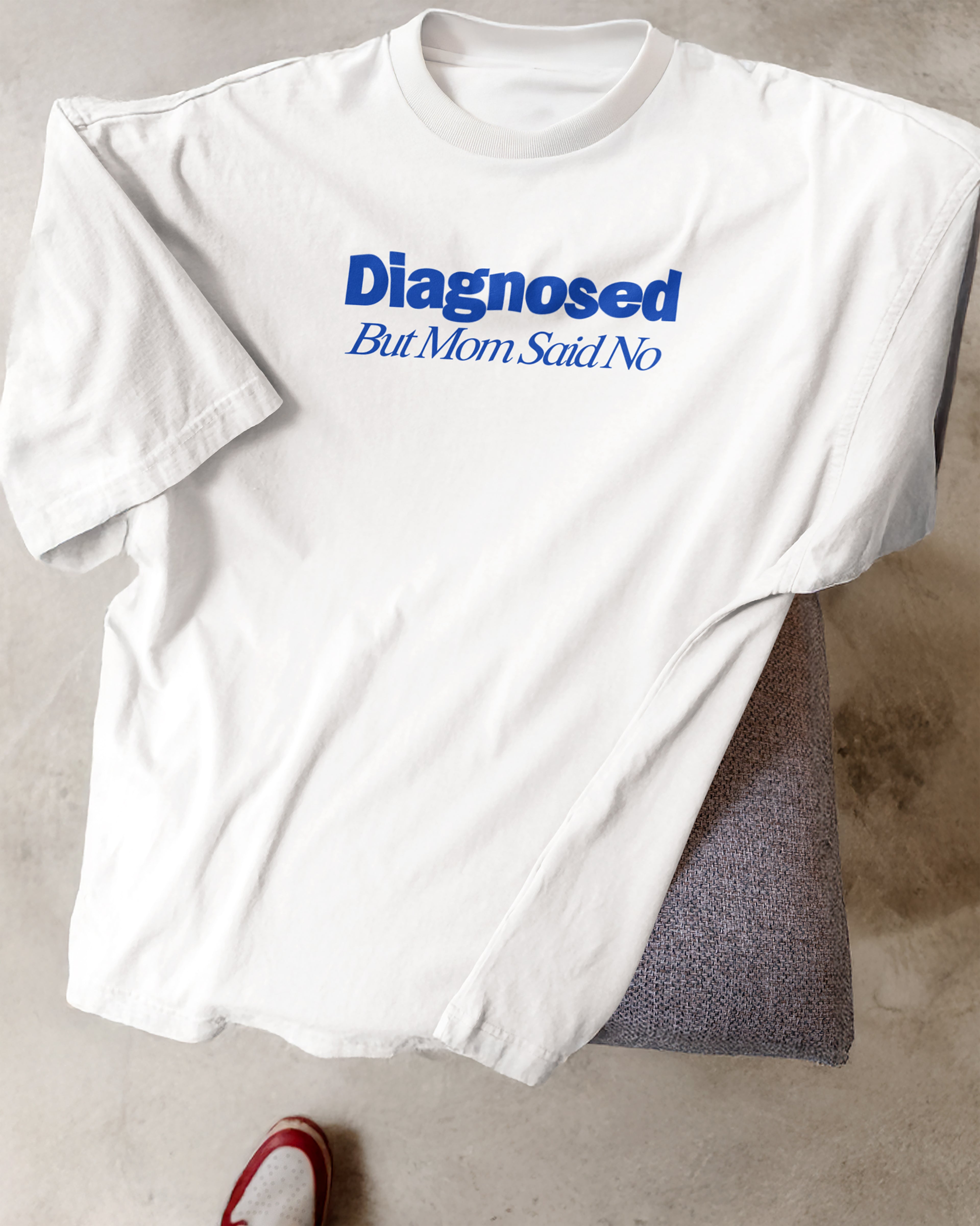 White t-shirt with blue text 'Diagnosed But Mom Said No' on a concrete floor.