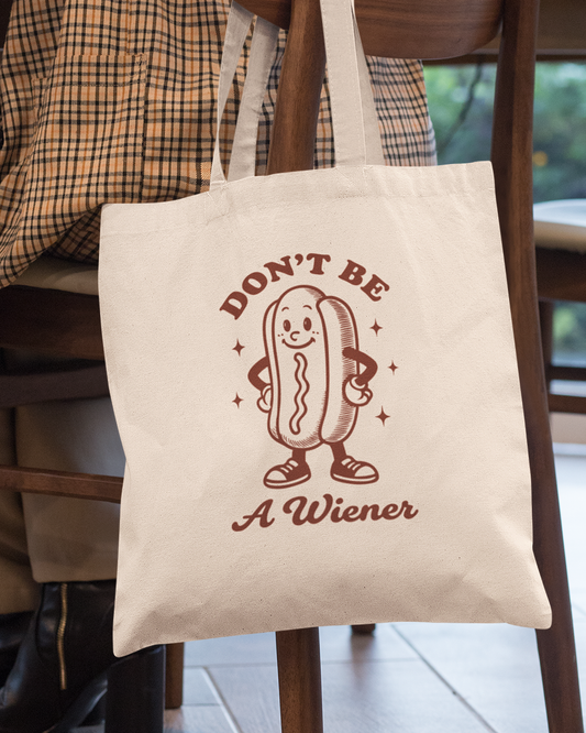 Tote bag with a cartoon hot dog and text 'Don't Be A Wiener' hanging on a chair.