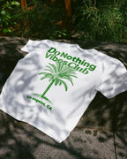 White t-shirt with green palm tree and 'do nothing vibes club' text on a stone surface with plants in the background