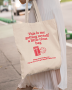 Tote bag with "this is my getting myself a little treat bag" text and graphics held by a person on a sidewalk.