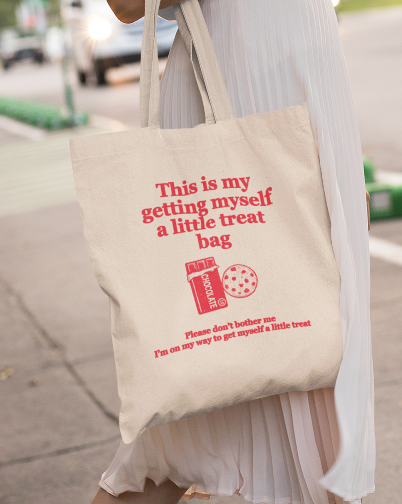 Tote bag with "this is my getting myself a little treat bag" text and graphics held by a person on a sidewalk.