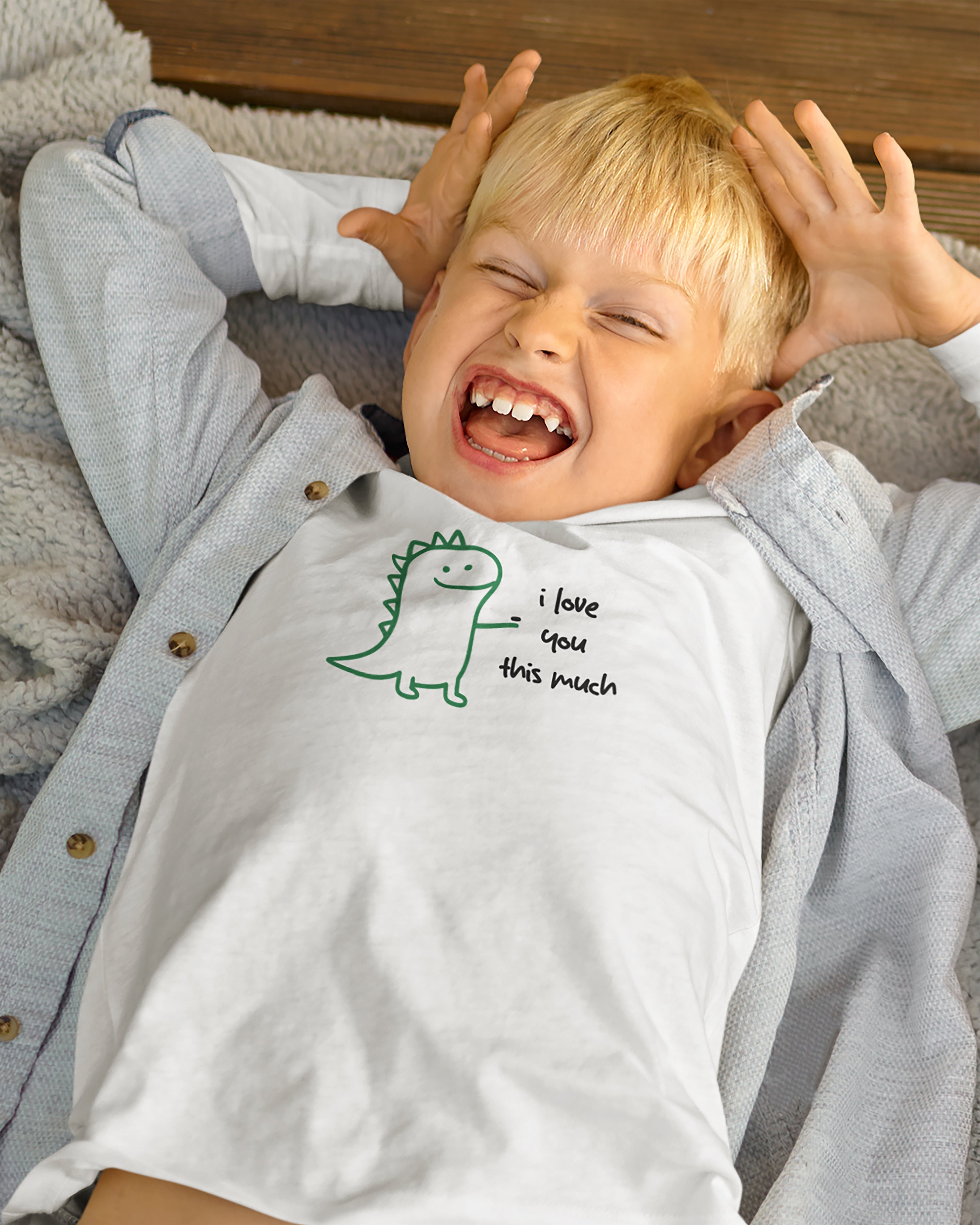 Child wearing a shirt with a dinosaur design and i love you this much text, lying on a bed.