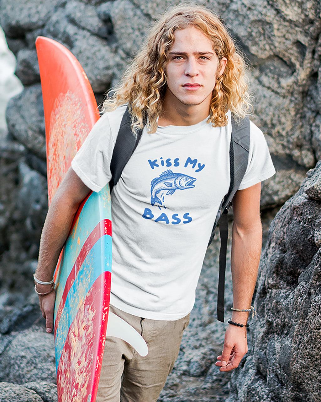 Person holding a surfboard with 'Kiss My Bass' t-shirt on rocky background