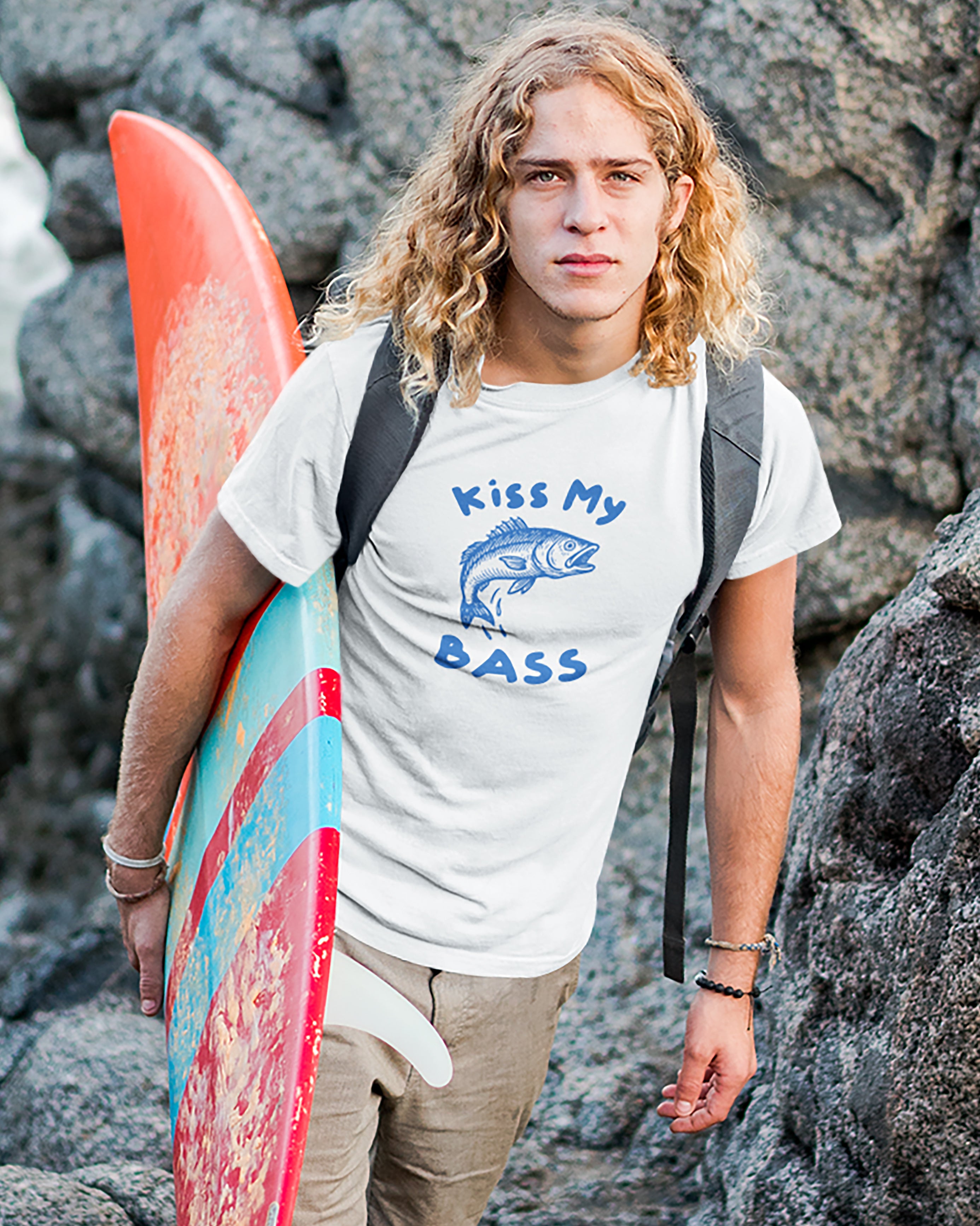 Person holding a surfboard with 'Kiss My Bass' t-shirt on rocky background