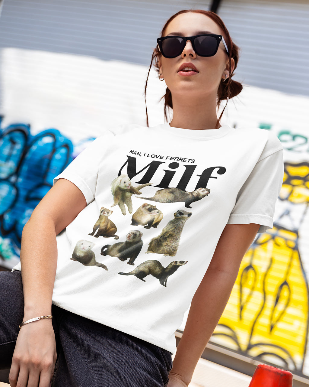 Person wearing a white t-shirt with ferret illustrations and "man, i love fettets" text, sitting in front of a colorful mural.