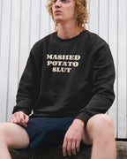 Person wearing a black sweatshirt with 'MASHED POTATO SLUT' text, sitting against a white wall.