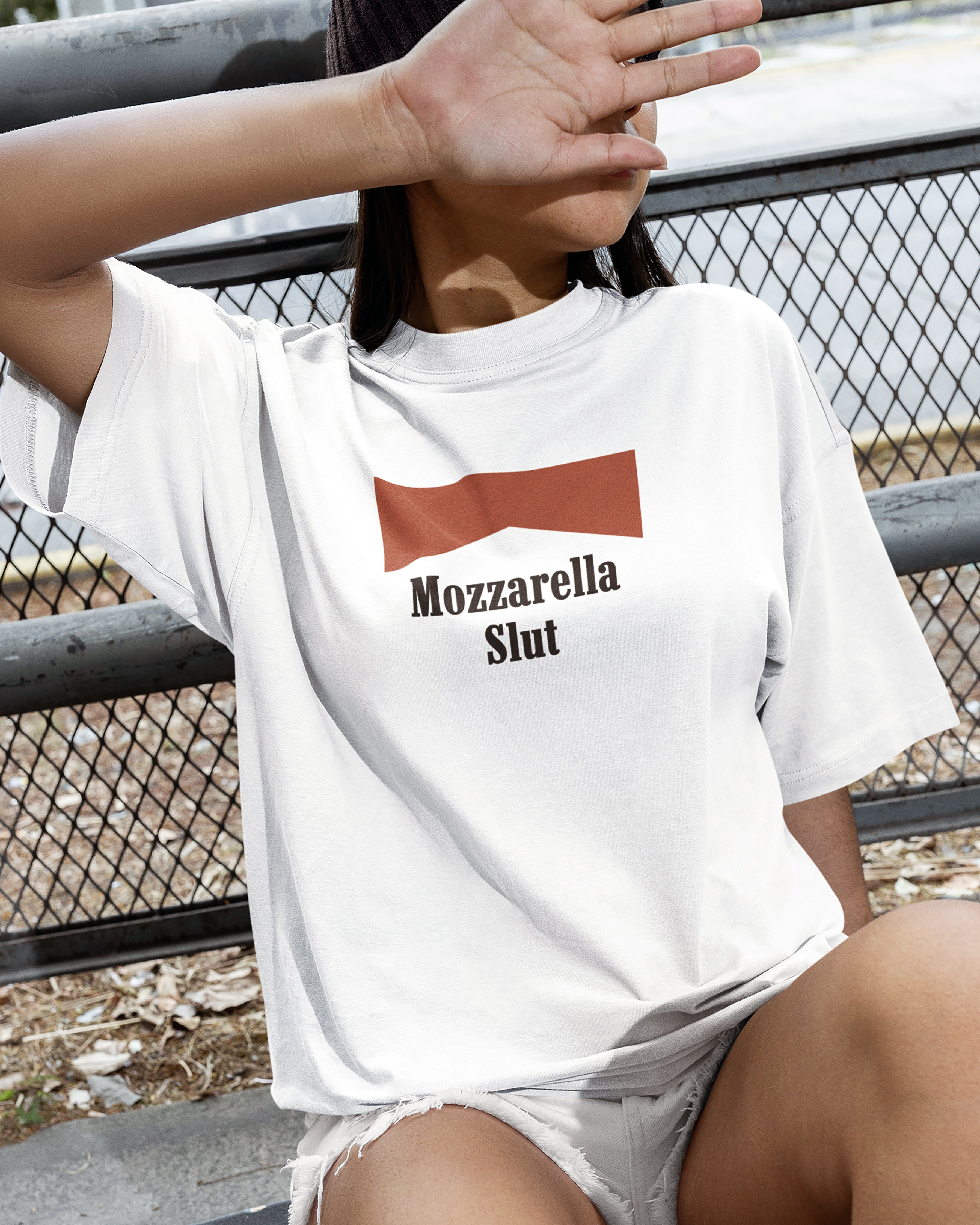 Person wearing a white t-shirt with 'Mozzarella Slut' text and red graphic, sitting on a bench.