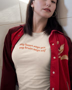 Woman wearing a red jacket and beige t-shirt with 'my heart says yes my brain says wtf' text, sitting against a light-colored wall.