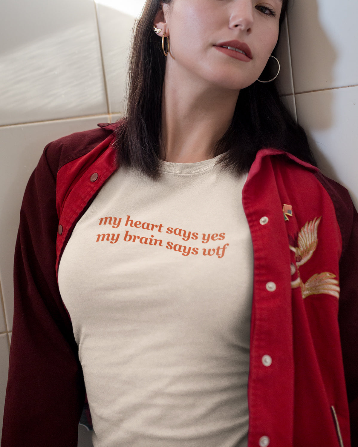 Woman wearing a red jacket and beige t-shirt with 'my heart says yes my brain says wtf' text, sitting against a light-colored wall.