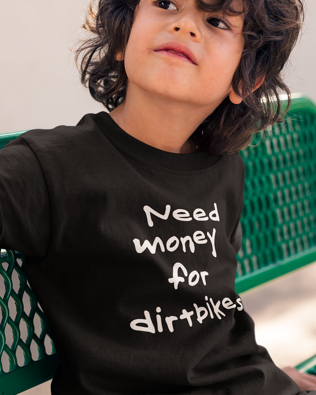 Child wearing a black t-shirt with need money for dirtbikes text sitting on a green bench.