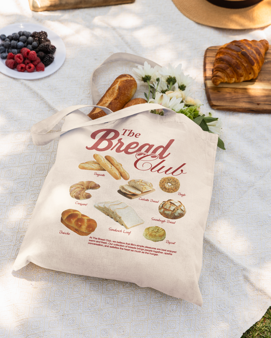 Picnic setup with 'The Bread Club' tote bag, bread, fruit, and wine on a blanket outdoors.