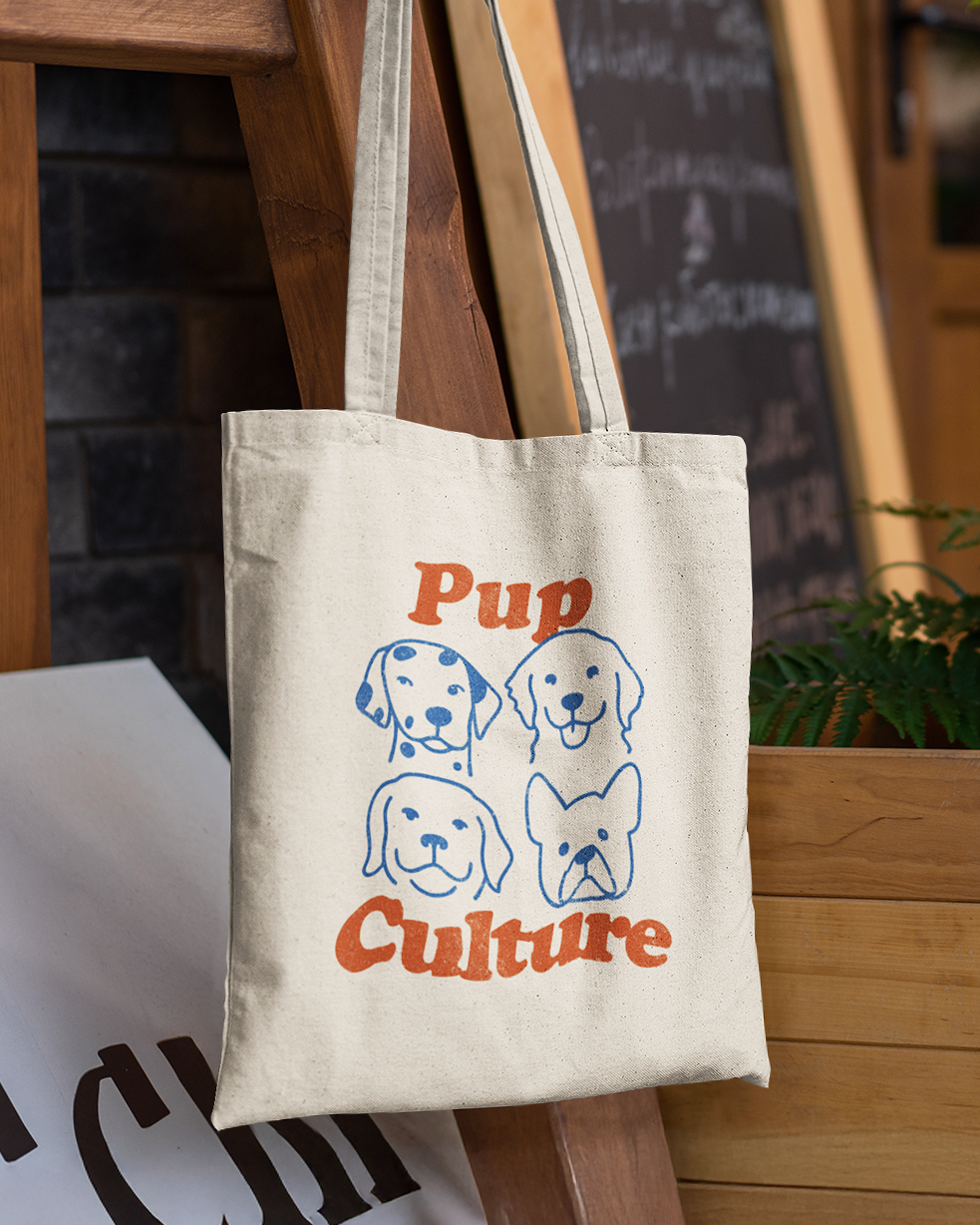 Tote bag with dog illustrations and 'Pup Culture' text hanging on a wooden surface.