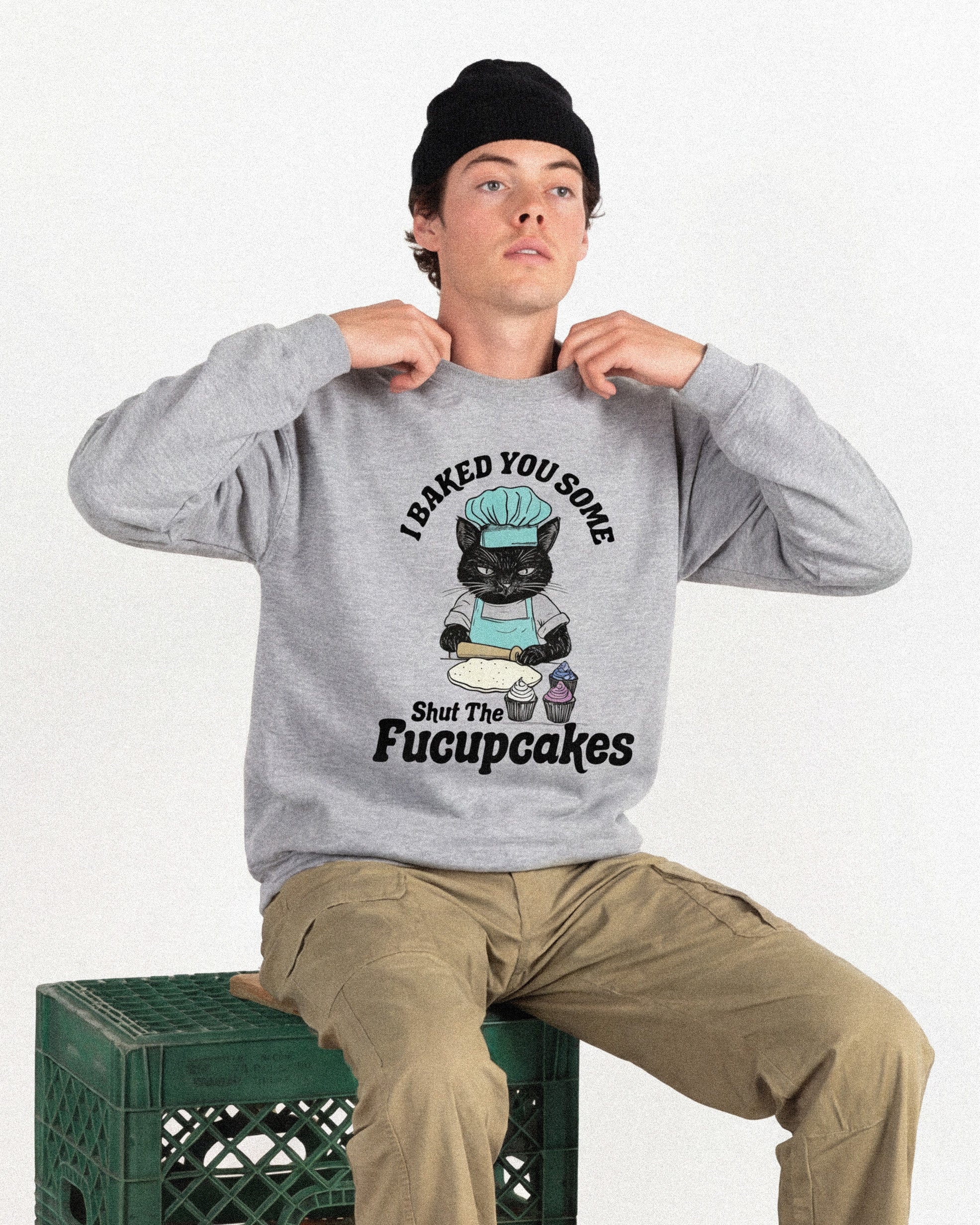 Person wearing a gray sweatshirt with a graphic design and text, sitting on a green crate against a white background.