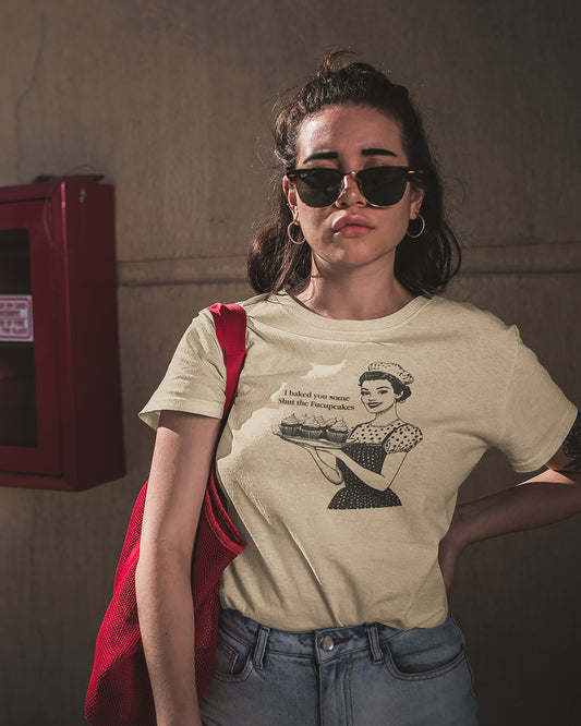 Woman wearing a beige t-shirt with a 'I Baked You Some Shut The Fucupcakes' graphic design, standing against a plain wall.