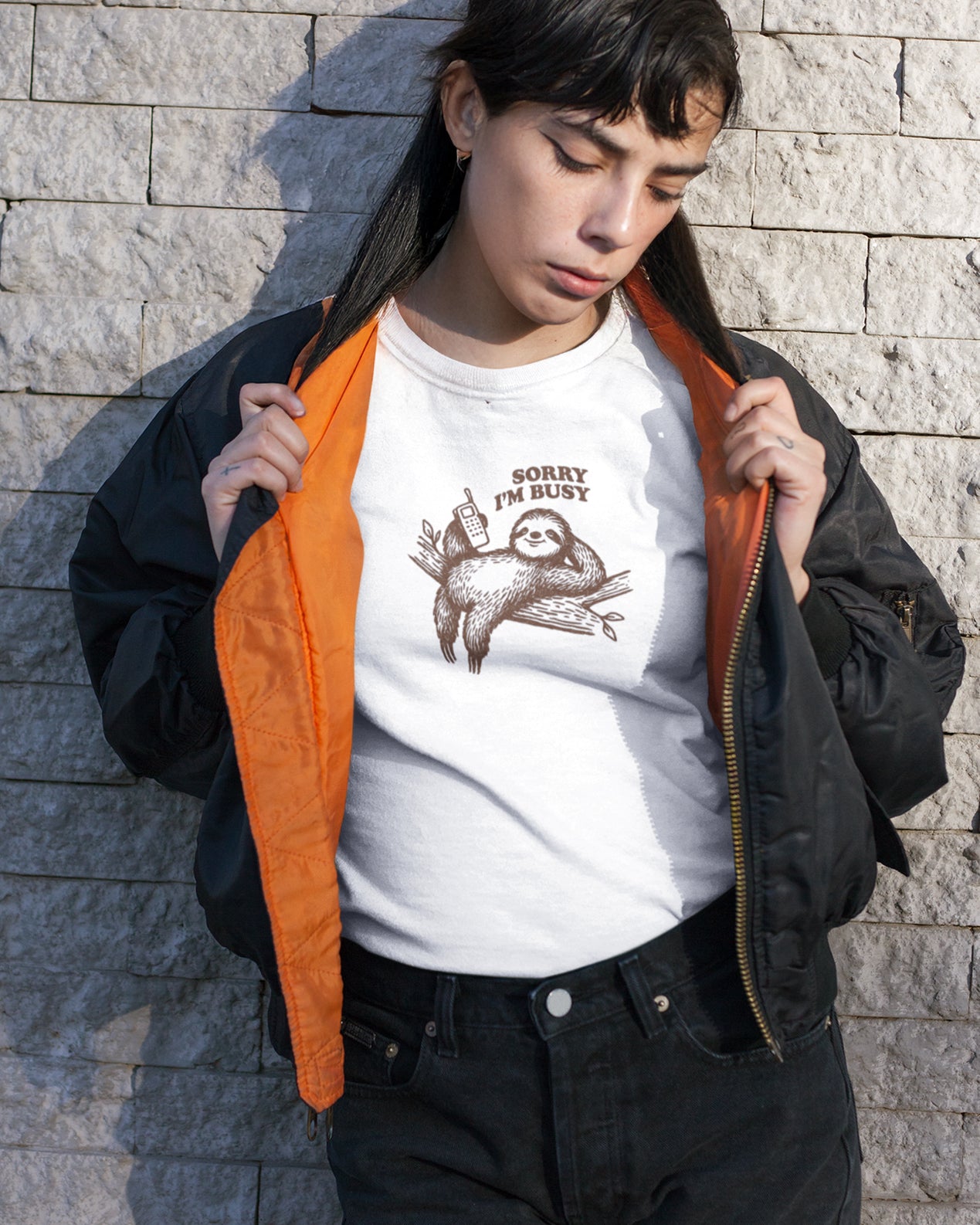Person wearing a white t-shirt with a sloth 'sorry i'm busy' graphic and text, standing against a stone wall.