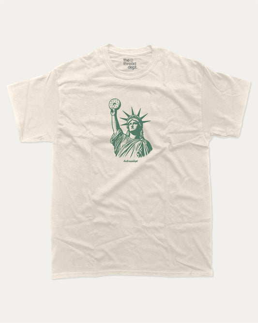 Statue of Liberty Bagle graphic tee with bold and humorous print, funny new york city graphic. Perfect for adults and gift-giving. Cream shirt.