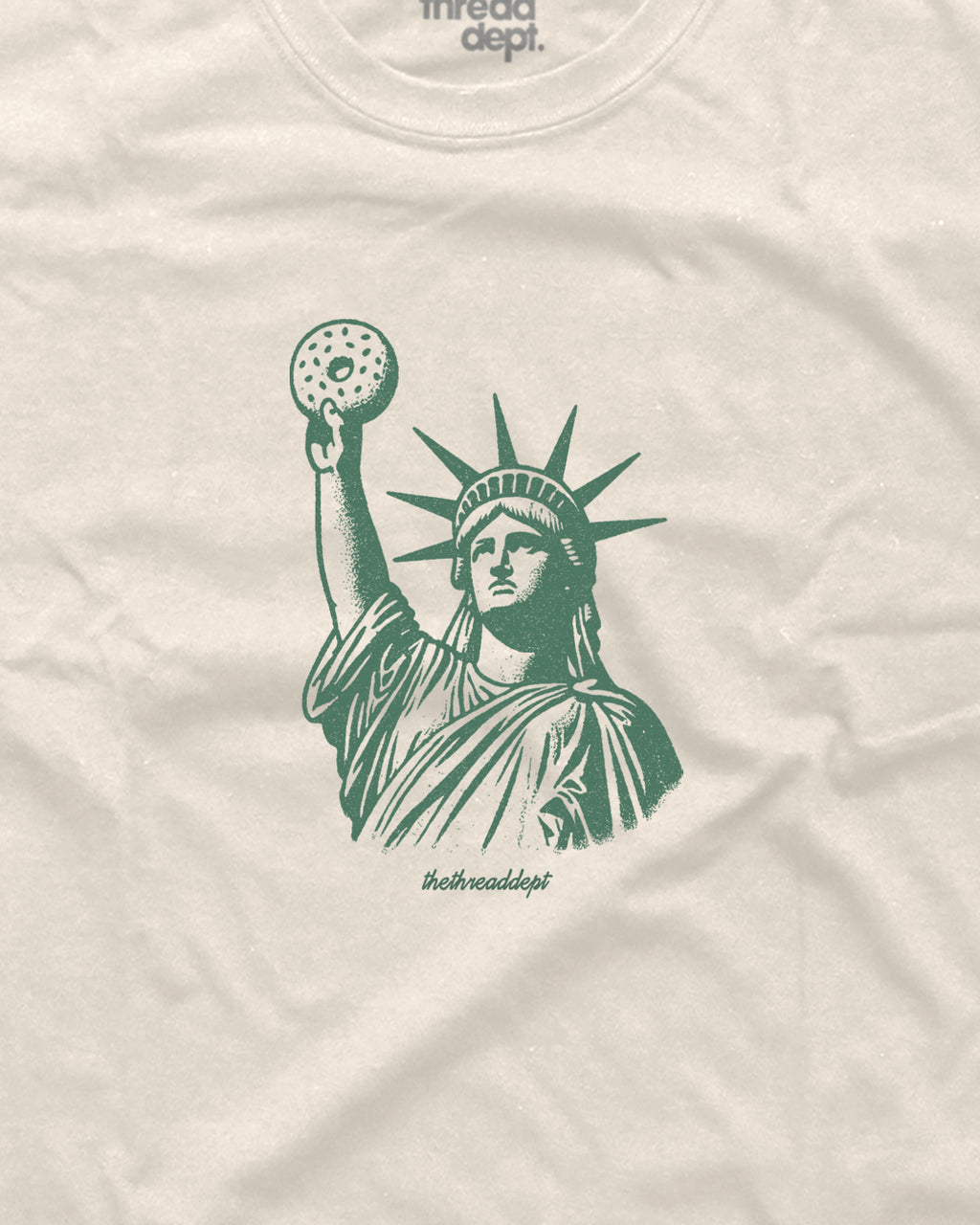 Beige t-shirt with green graphic of the Statue of Liberty holding a bagle.