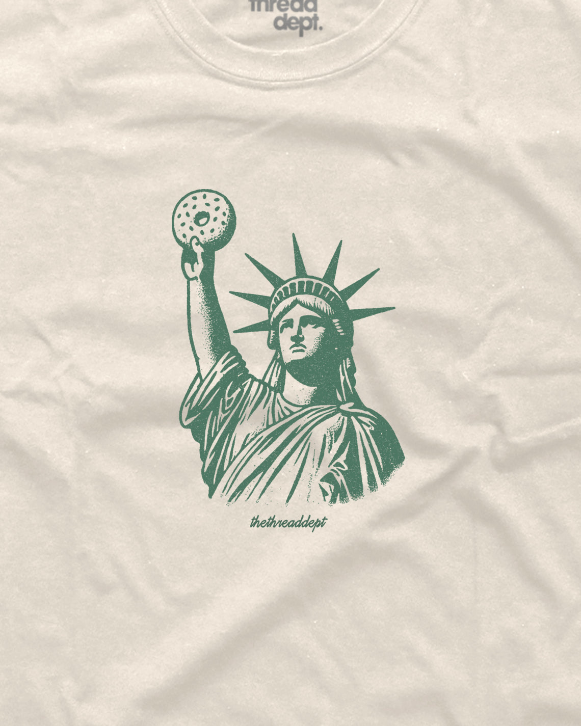 Beige t-shirt with green graphic of the Statue of Liberty holding a bagle.