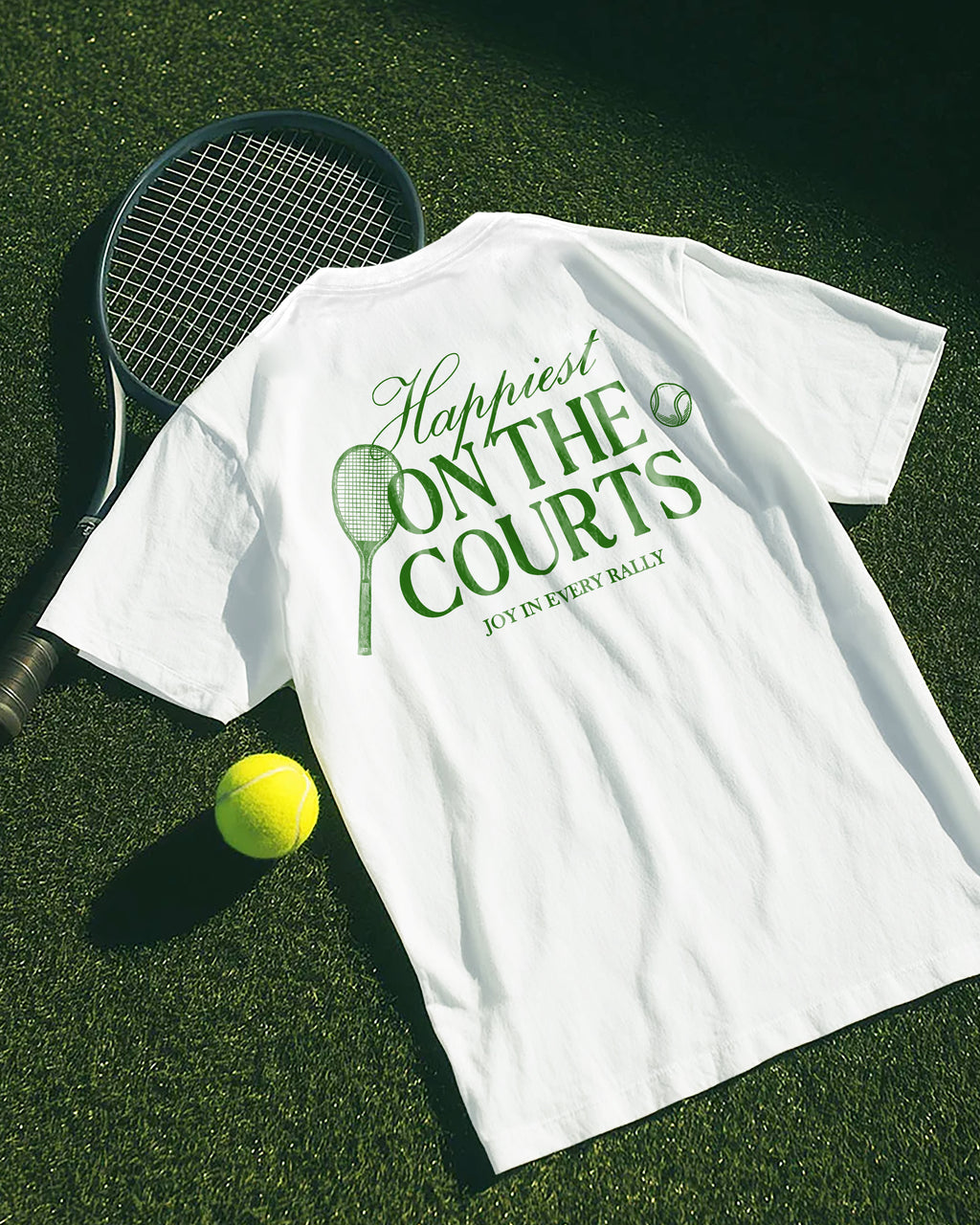 White t-shirt with 'Happiest on the Courts' text, tennis racket, and ball on grass