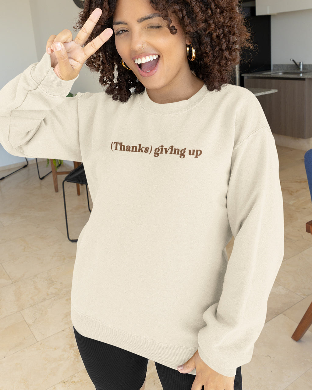 Person wearing a beige sweatshirt with Thanks Giving Up graphic, making a peace sign indoors.