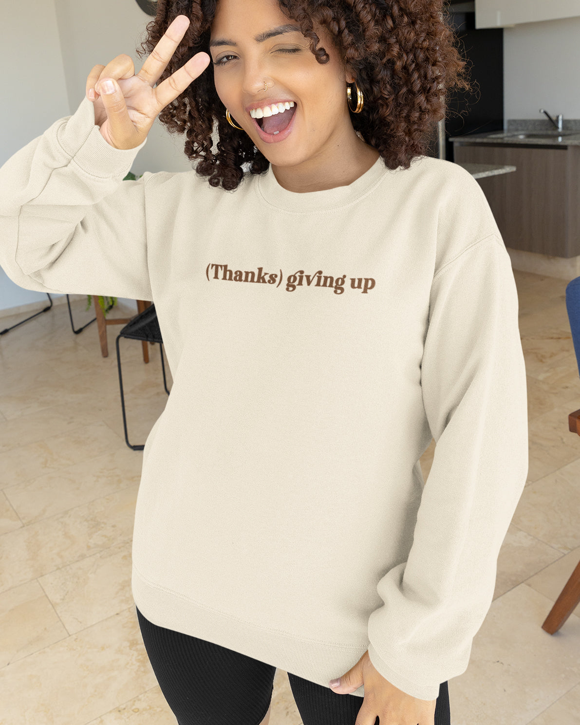Person wearing a beige sweatshirt with Thanks Giving Up graphic, making a peace sign indoors.