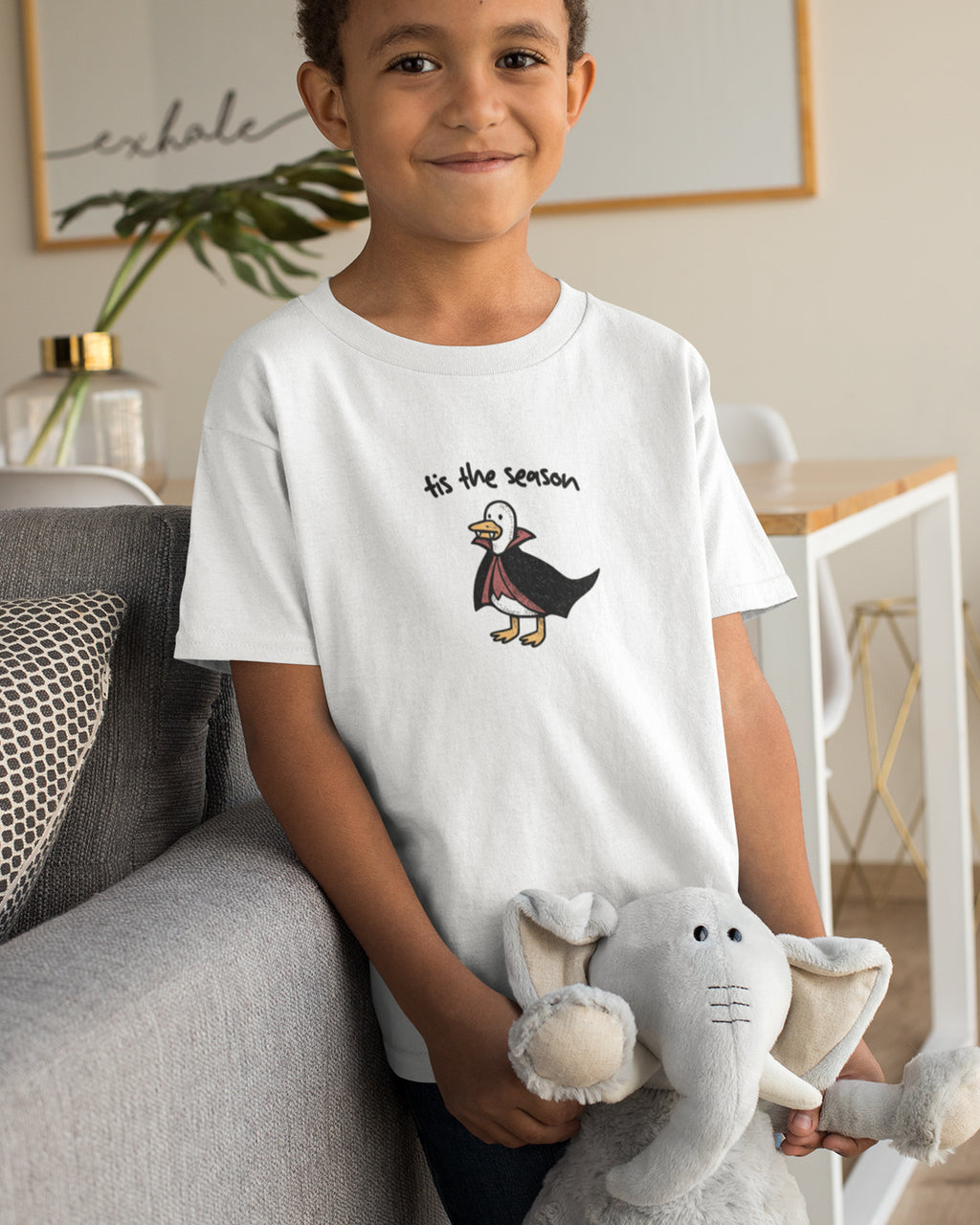 Child wearing a white t-shirt with a graphic design, sitting on a couch holding a stuffed elephant toy.