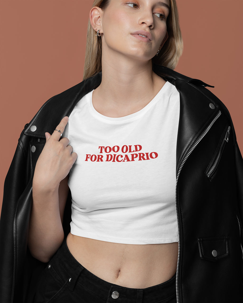 Person wearing a white crop top with 'Too Old for DiCaprio' text, standing against a brown background.