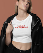 Person wearing a white crop top with 'Too Old for DiCaprio' text, standing against a brown background.