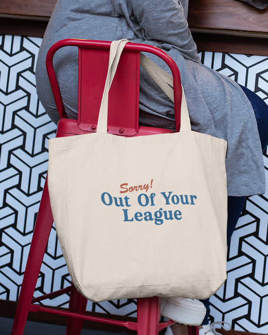 Beige tote bag with 'Sorry! Out Of Your League' text on a red chair, person wearing gray sweatshirt and blue pants.