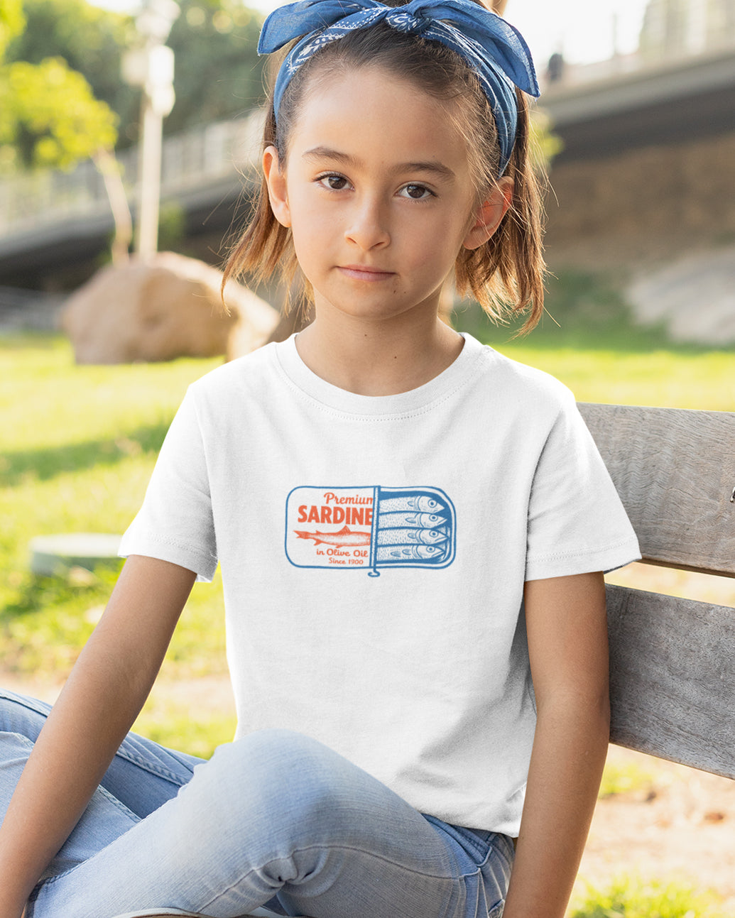 Young girl sitting on a bench wearing a white t-shirt with a Tinned Sardines graphic design.