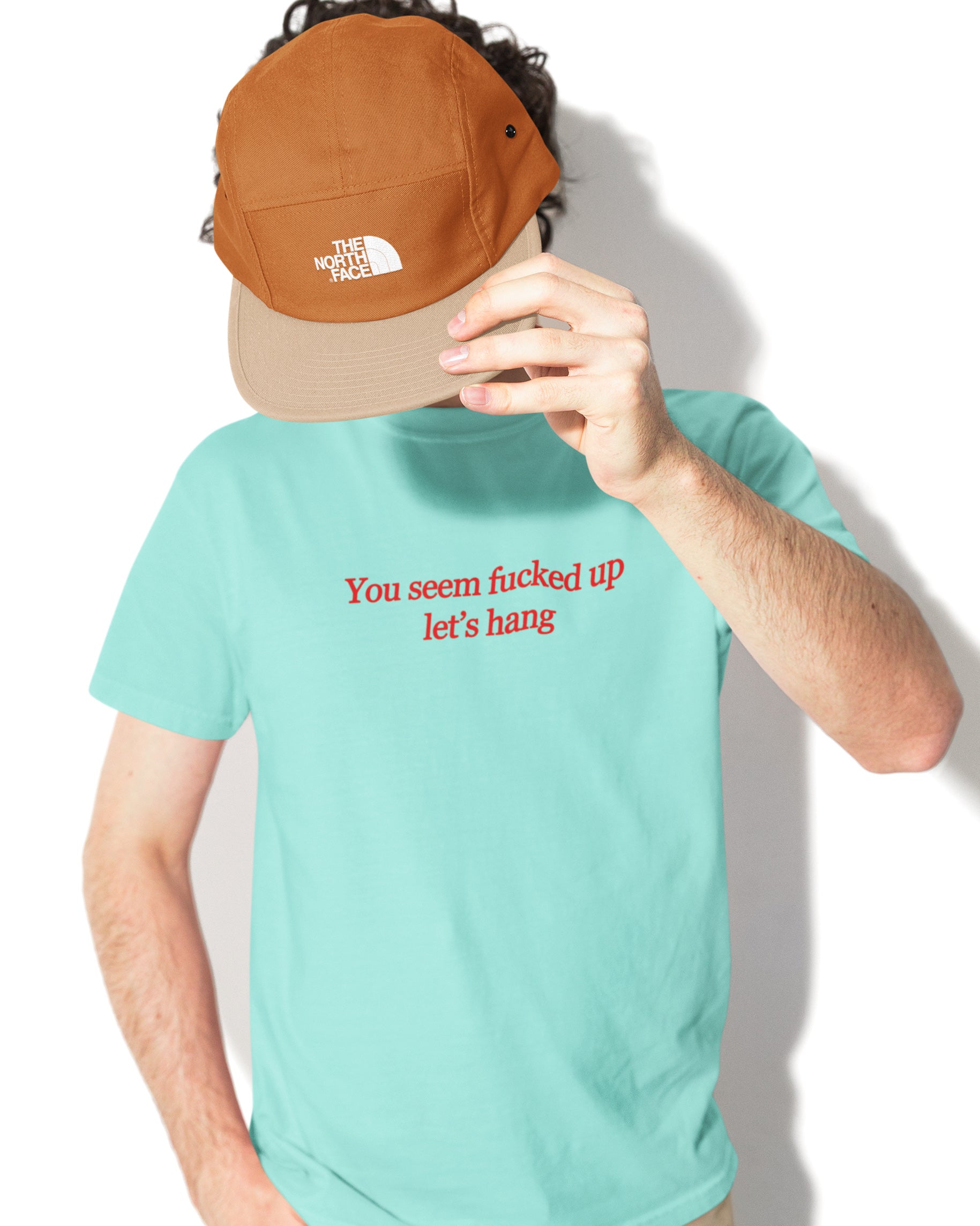Person wearing a turquoise t-shirt with "you seem fucked up let's hang" text and a brown cap held up to the camera.